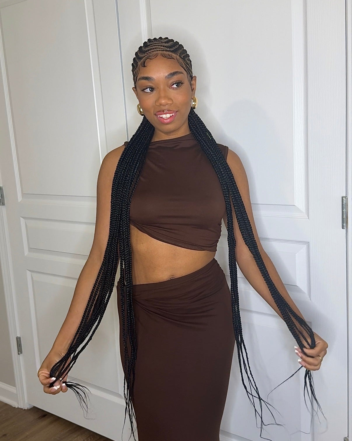 Woman in a brown outfit holding long braided hair extensions against a white door.